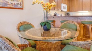 Een eetgedeelte met een tafel met glazen blad, omringd door rieten stoelen, een potplant en een keuken op de achtergrond.
