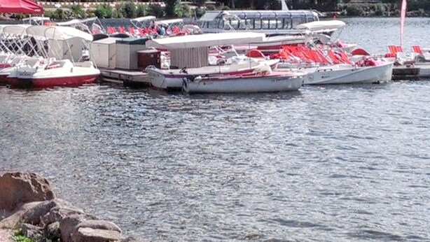 Een pier aan het meer met boten, een gebouw met rode daken en een beboste heuvel op de achtergrond.
