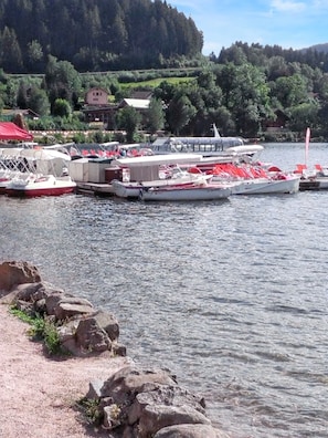 A lakeside dock with boats, a red-roofed building, and a forested hillside in the background.