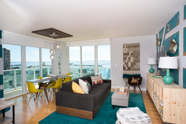 A modern living room with a large window, a dark grey sofa, yellow chairs, and a teal rug.