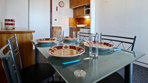 A dining area with a glass table set for four, surrounded by chairs. A kitchen is visible in the background.