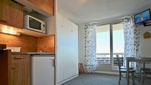 A compact kitchen with a microwave, a small refrigerator, and wooden cabinets. There is a dining area with a table and chairs, and a balcony with curtains.