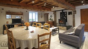 A rustic interior with exposed wooden beams, a dining area with a table and chairs, a sofa, a fireplace, and a television. - Large stone house with garden near the beach (Héauville)