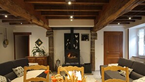 A cozy living room with a fireplace, wooden beams, a dining table with chairs, and a sofa. - Large stone house with garden near the beach (Héauville)