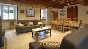A cozy living room with a sofa, a dining table with chairs, a wooden ceiling, and a clock on the wall. - Large stone house with garden near the beach (Héauville)