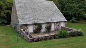 A stone cottage with a steeply pitched roof, surrounded by a garden with flowering plants and a grassy lawn. - Large stone house with garden near the beach (Héauville)