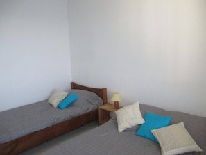 Quarto pequeno e simples com duas camas de solteiro, cada uma com travesseiro e mesa de cabeceira com abajur.
