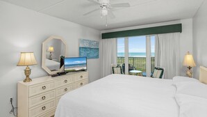 A hotel room with a large bed, a ceiling fan, a television, a dresser, a mirror, a window with curtains, and a view of the ocean.
