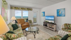 A living room with a sofa, armchair, and a glass coffee table. There is a television on a stand and a bookshelf.