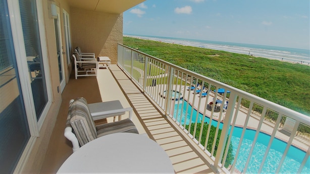 A balcony with a view of the beach, a pool, and outdoor seating.