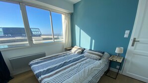 Un dormitorio con cama, mesita de noche, lámpara, radiador y ventana con vista a un edificio y una cerca.