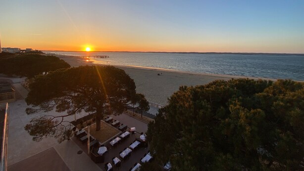 Vista al atardecer desde una terraza con vistas a una playa y un muelle, zona de comedor con mesas y sillas, y una hilera de árboles.