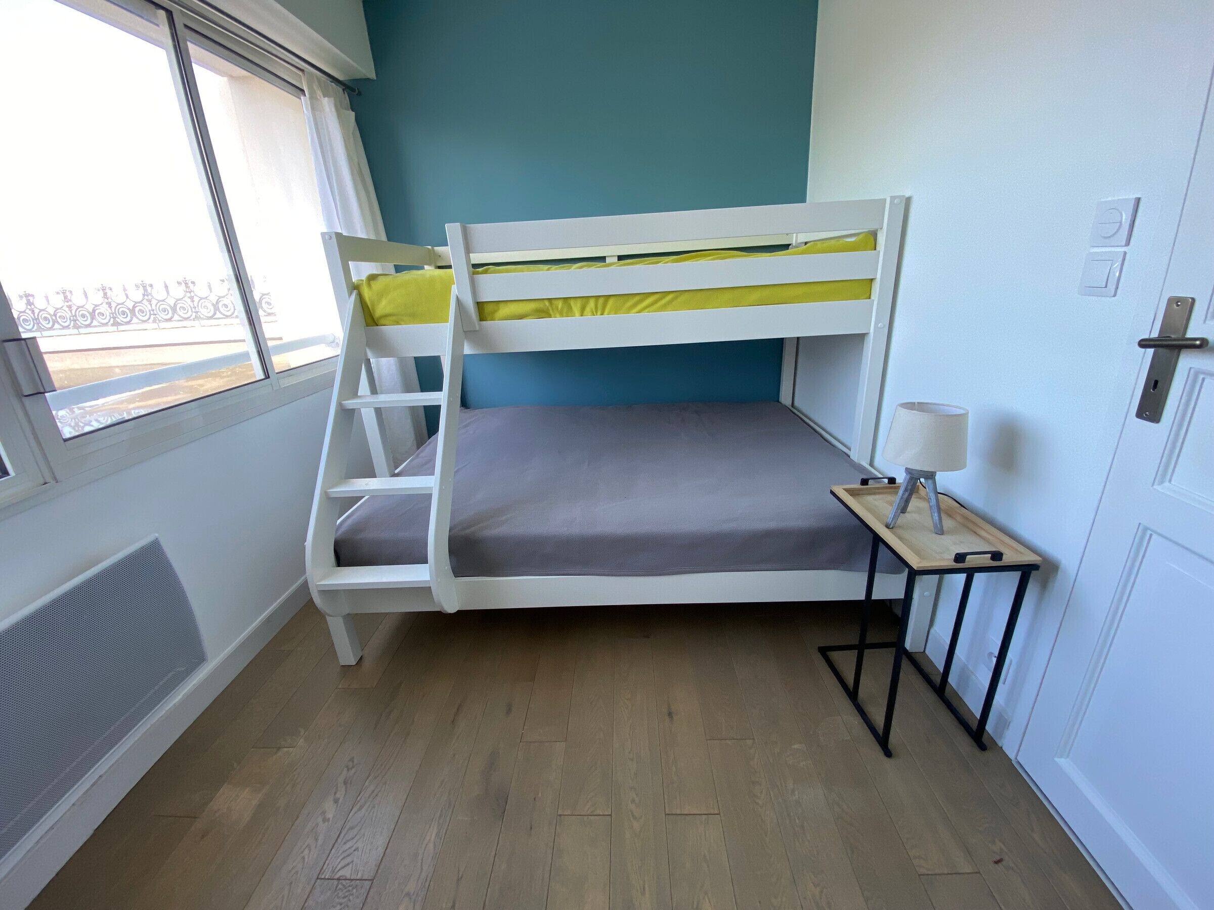 A bunk bed room with a window, a small table with a lamp, and a door.