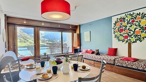 A room with a dining table set for breakfast, a sofa with a colorful apple tree mural, and a view of a mountainous landscape through large windows. - Tignes, 6 pers, 55m², balcony, parking included (Tignes)