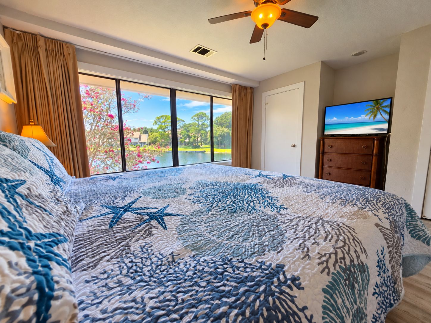 A bedroom with a large bed, a ceiling fan, a television, and a view of a lake.