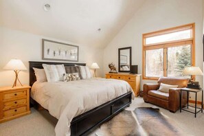 Un dormitorio con cama, mesitas de noche, una silla, una ventana y obras de arte en las paredes.