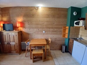 A compact kitchen with wooden cabinets, a dining table with chairs, a microwave, and a trash bin.