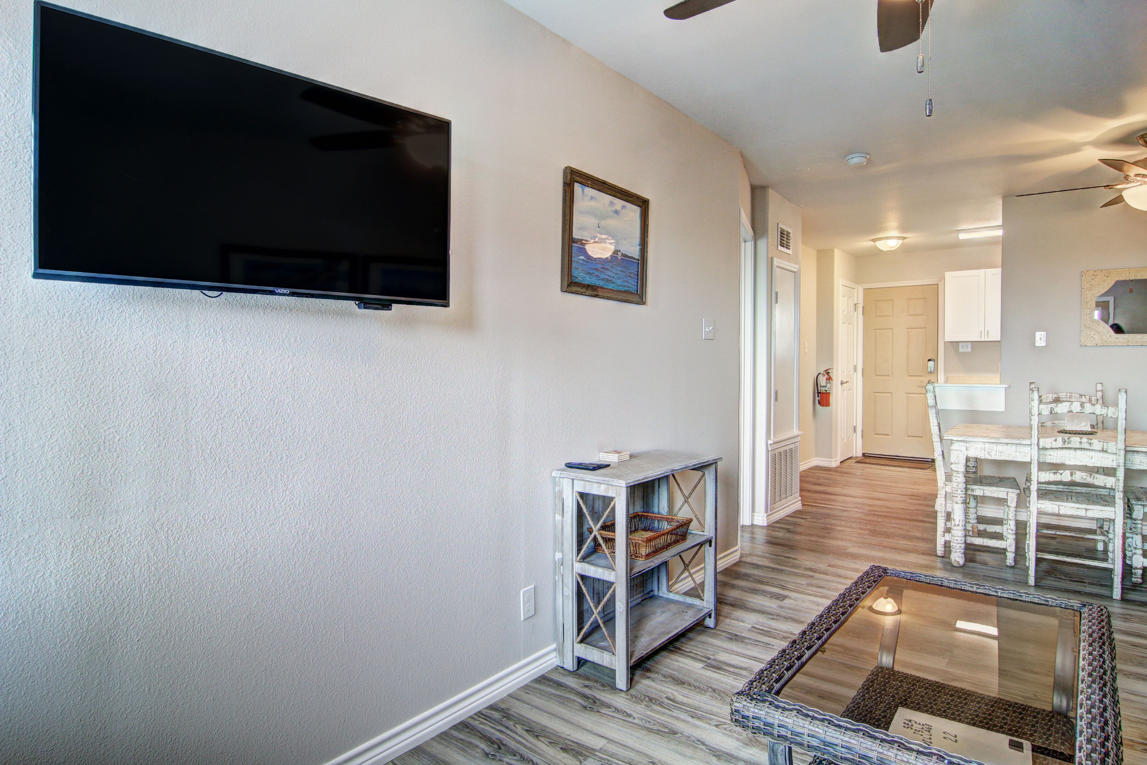 A living room with a flat-screen TV, a small table with baskets, a glass coffee table, and a dining area with white chairs.