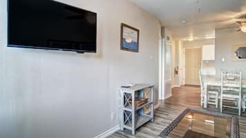 A living room with a flat-screen TV, a small table with baskets, a glass coffee table, and a dining area with white chairs.