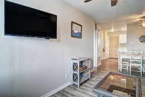 A living room with a flat-screen TV, a small table with baskets, a glass coffee table, and a dining area with white chairs. - Beautiful Gulf Views at Sea Isle Village! (Port Aransas)