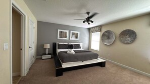 A bedroom with a bed, a ceiling fan, two wall-mounted picture frames, a window with blinds, and a decorative wall plate. - Private Pool & Spa | No Rear Neighbors | Walk to Clubhouse | 6BR Near Disney (Kissimmee)