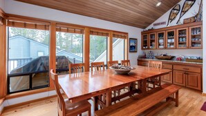 A spacious dining area with a large wooden table and benches, a kitchen with wooden cabinets, and large windows offering views of the outdoors. - CHER06 - Great for Groups, A/C, Hot Tub (Sunriver)