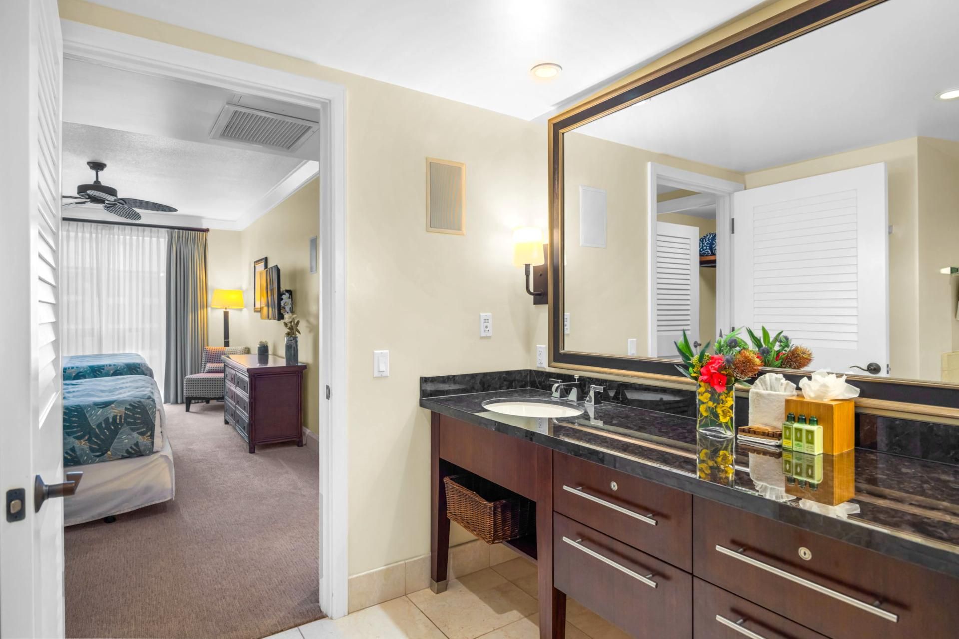 A hotel bathroom with a large mirror, a sink, and a vanity. There is a ceiling fan above the bed, and a chair with a lamp in the adjacent room.