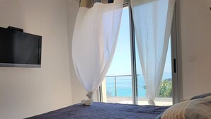 Quarto com cama, TV na parede e vista para o mar através de uma varanda.