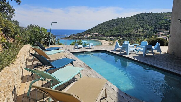Uma área de piscina com espreguiçadeiras e vista para o mar e colinas.