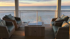 A covered outdoor seating area with wicker furniture and a wooden table, overlooking a beach with a wooden deck and a railing.