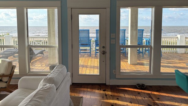 A room with a white couch, a wooden floor, and a view of the beach through large windows. - 2 Story Upscale Beachfront House With unobstructed Beach Views from every room. (Crystal Beach)