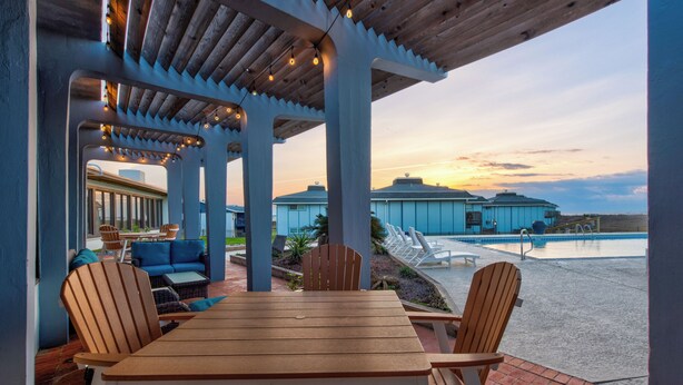Un espacio de estar al aire libre techado, con una mesa y sillas de madera, con vistas a una piscina y un edificio residencial.