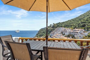 Un balcon avec des fauteuils et une table en osier, offrant une vue sur une ville côtière et la mer.