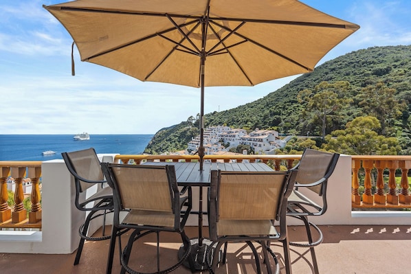 A terrace with a table and chairs, an umbrella, and a view of the sea and a hillside town.