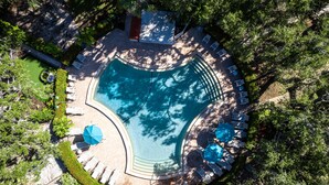 An aerial view of a swimming pool surrounded by lounge chairs and umbrellas, with a small deck area and dense foliage in the background.