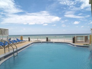 A pool with a view of the beach and ocean, surrounded by lounge chairs and a fence.
