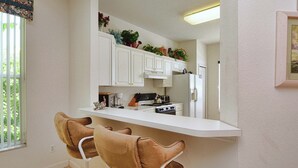 Una cocina con gabinetes blancos, una encimera blanca, dos taburetes marrones y un refrigerador.
