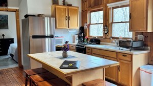 A kitchen with wooden cabinets, a stainless steel refrigerator, wooden stools, and a large island. - Peace-in-the-Forest Retreat - Singles, Couples, Artists, Forest Lovers (Pickens)