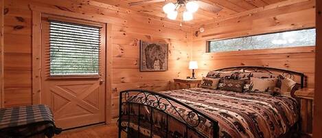 A wooden cabin bedroom with a bed, a ceiling fan, a window, and a door.