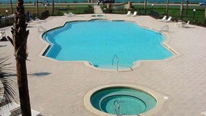 Uma piscina ampla e de formato irregular com uma banheira de hidromassagem circular menor no centro, rodeada por espreguiçadeiras e palmeiras.