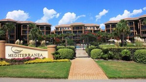 Entrada de hotel com uma placa indicando "Mediterranea", rodeada por paisagismo bem cuidado e palmeiras.