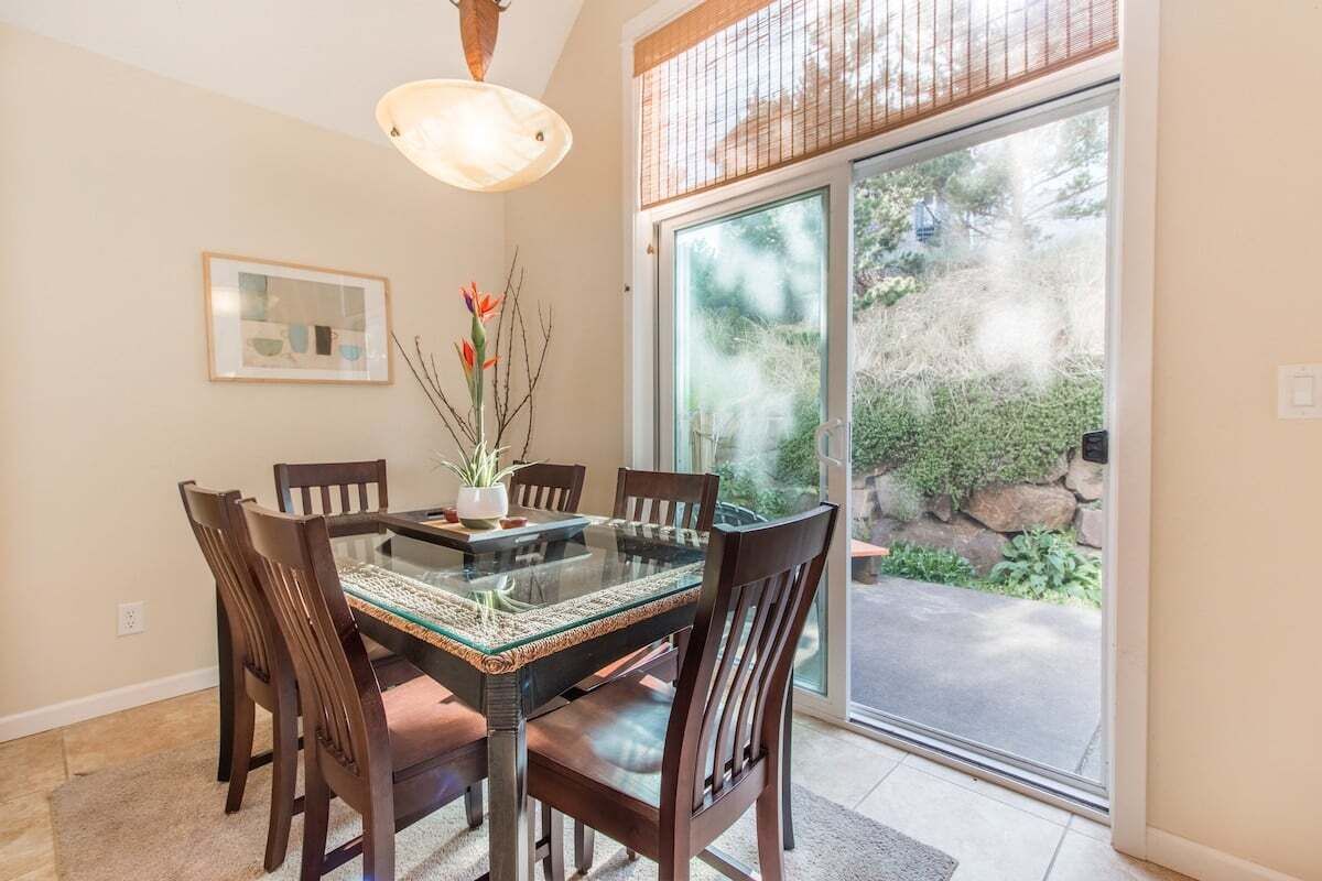A dining area with a glass-top table, wooden chairs, a hanging light fixture, a framed picture on the wall, and a view of a garden through a glass door.