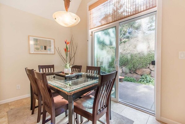 A dining area with a glass-top table, wooden chairs, a hanging light fixture, a framed picture on the wall, and a view of a garden through a glass door.