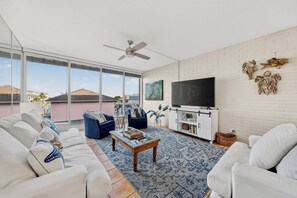 A modern living room with a large TV, white sofas, a coffee table, and a view of the city. - This spacious 2/2 is located right in the heart of Port A! Private fishing pier! (Port Aransas)