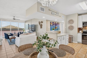 A modern living room with a white sofa, a dining area with wicker chairs, a kitchen with white cabinets, and a large window overlooking a hilly landscape. - This spacious 2/2 is located right in the heart of Port A! Private fishing pier! (Port Aransas)