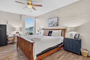 A bedroom with a wooden bed, white brick walls, a ceiling fan, and a window with a view of a blue house. - This spacious 2/2 is located right in the heart of Port A! Private fishing pier! (Port Aransas)