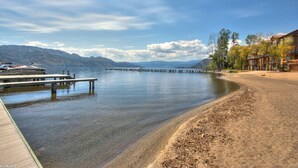 A lakeside view with a wooden dock extending into calm water, surrounded by mountains and a building with balconies. - Barona Beach Lakefront Condo (West Kelowna)