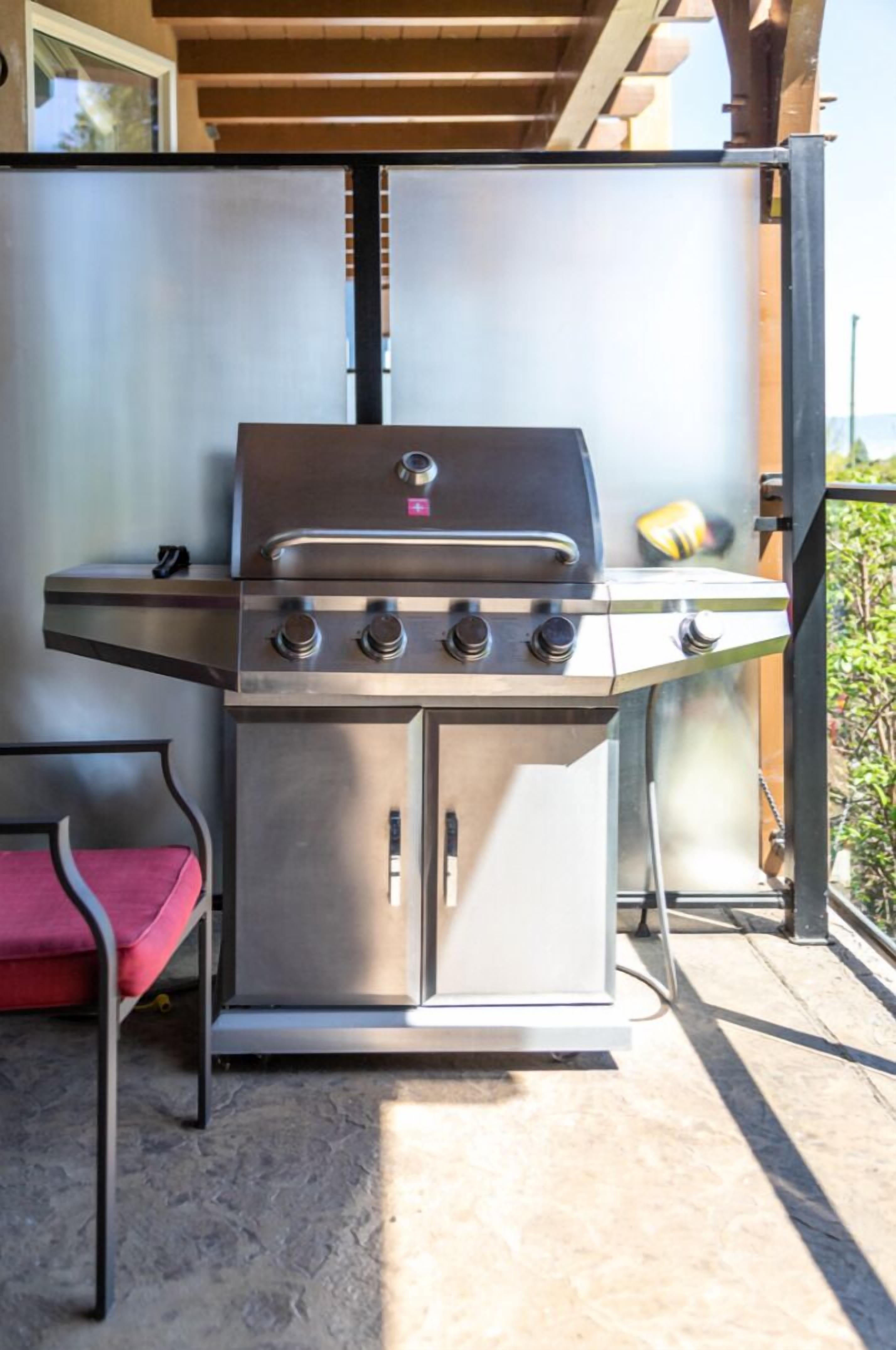 En grill i rustfrit stål med fire brændere, en rød polstret stol og en balkon med udsigt over grønne områder.