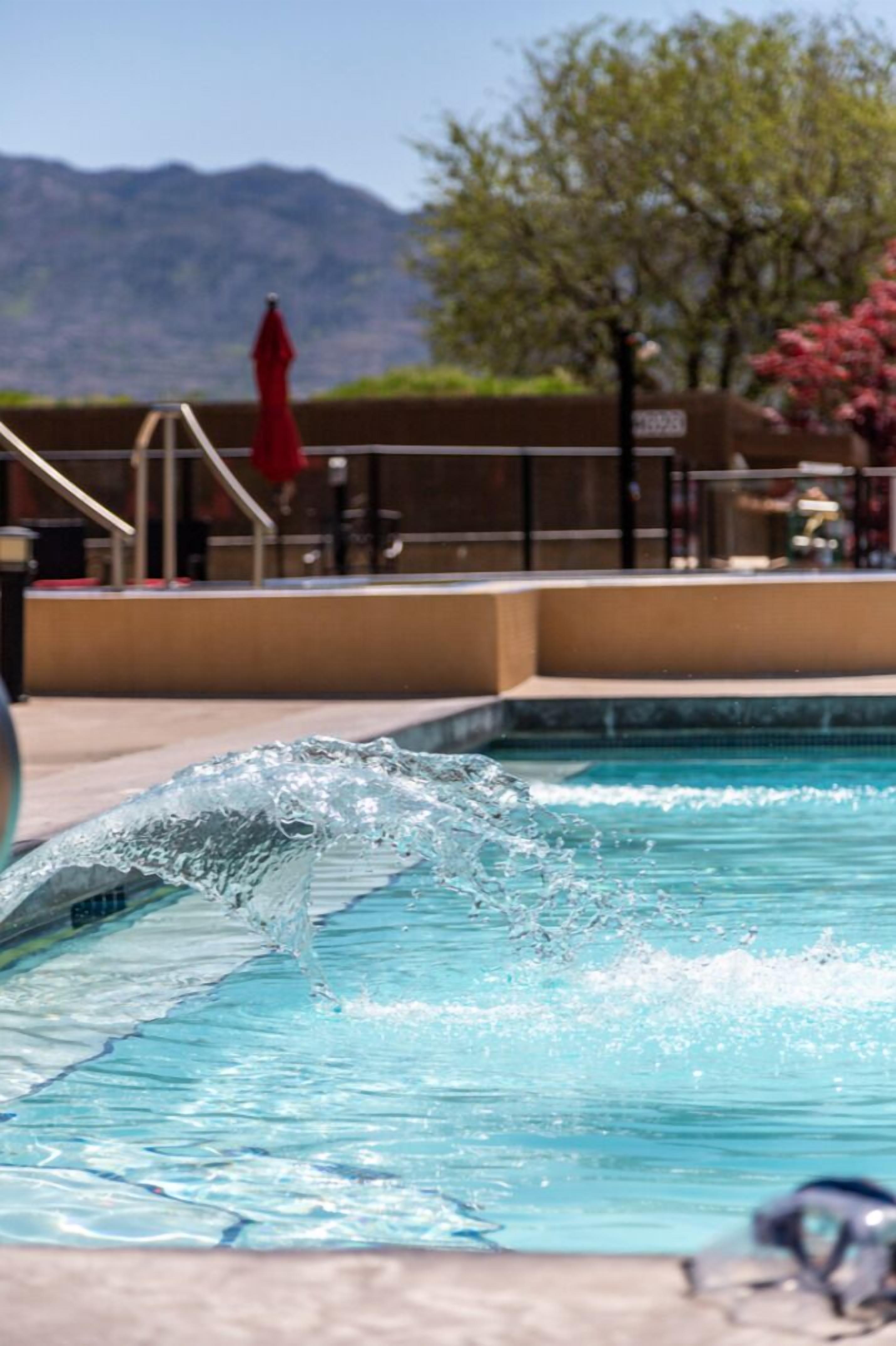Ein Schwimmbecken mit spritzendem Wasser, einem roten Sonnenschirm und einem Berg im Hintergrund.