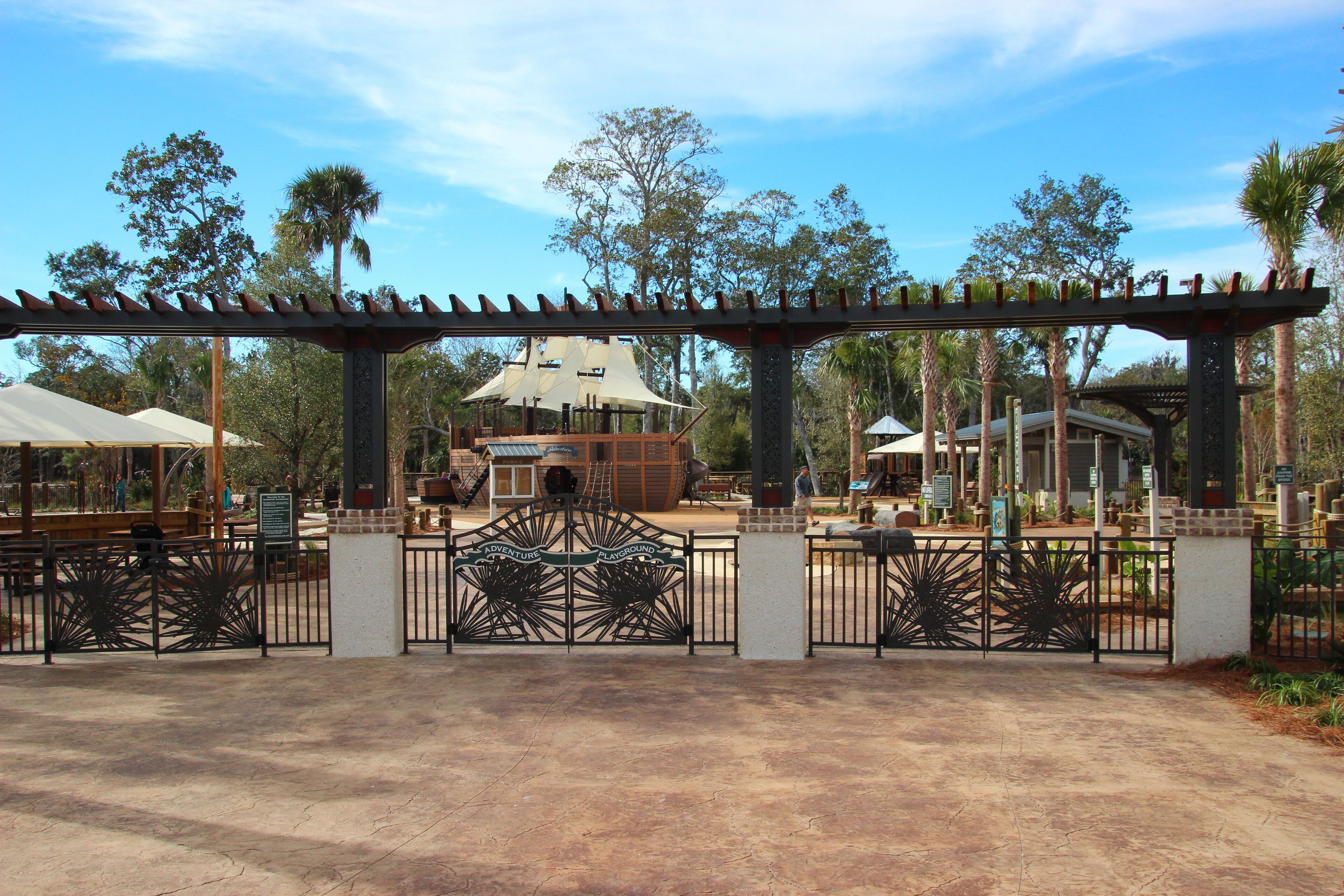 A park entrance with a pergola, a play area, and a large tree in the background.